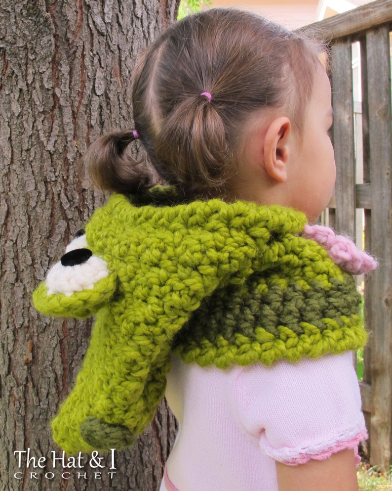 May include: A green and brown crocheted hooded scarf with a frog face on the hood. The scarf is worn by a child with pigtails.