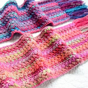 Pu&ograve; includere: Una sciarpa all'uncinetto colorata con un motivo testurizzato. La sciarpa &egrave; realizzata con una variet&agrave; di tonalit&agrave; di rosa, viola, blu e verde. La sciarpa &egrave; piegata a met&agrave; e le estremit&agrave; sono visibili nell'immagine. Il testo "The Hat & I Crochet" &egrave; visibile nell'angolo in alto a destra dell'immagine.