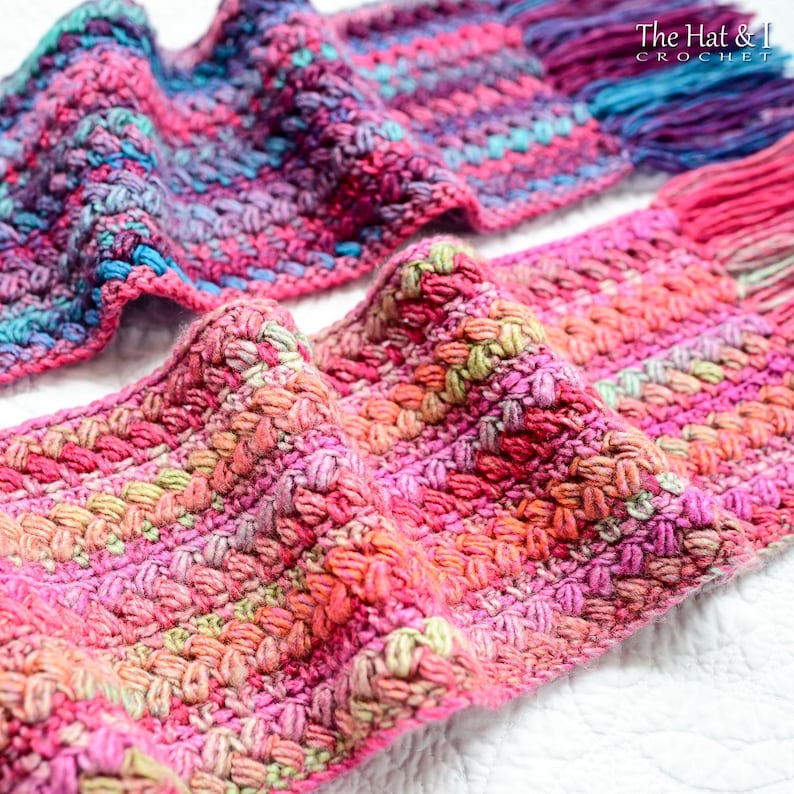 Pu&ograve; includere: Una sciarpa all'uncinetto colorata con un motivo testurizzato. La sciarpa &egrave; realizzata con una variet&agrave; di tonalit&agrave; di rosa, viola, blu e verde. La sciarpa &egrave; piegata a met&agrave; e le estremit&agrave; sono visibili nell'immagine. Il testo "The Hat & I Crochet" &egrave; visibile nell'angolo in alto a destra dell'immagine.