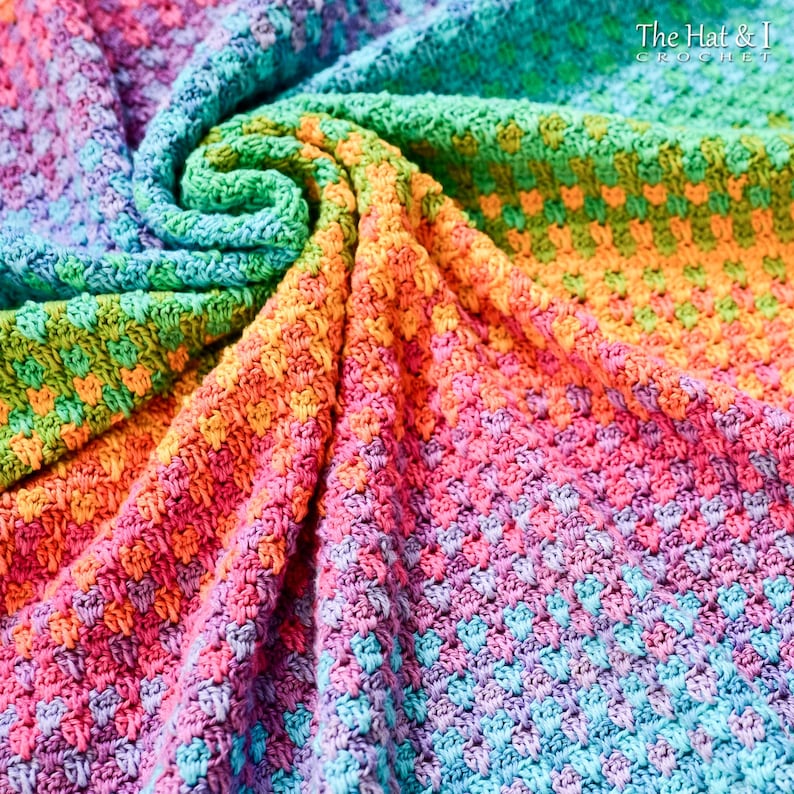 May include: A rainbow crochet blanket with a checkered pattern. The blanket is made with a variety of colors, including red, orange, yellow, green, blue, and purple. The blanket is folded in a spiral shape, showing off the different colors and textures. The text "The Hat & I Crochet" is visible in the upper right corner of the image.