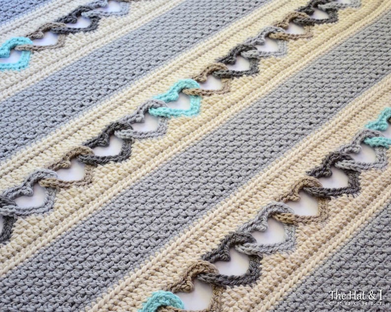 May include: A crocheted blanket with a striped pattern in shades of gray, beige, and teal. The blanket features a repeating heart design created with crocheted loops.