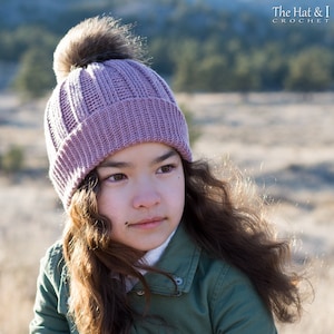 Pu&ograve; includere: Una giovane persona indossa un berretto di lana color malva con un pon pon marrone. Il berretto ha una trama a coste. La persona indossa una giacca verde e ha i capelli lunghi e castani.