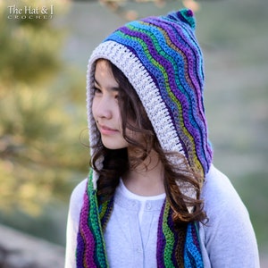 Pu&ograve; includere: Una sciarpa con cappuccio all'uncinetto colorata con un motivo ondulato in tonalit&agrave; di viola, blu, verde e bianco. La sciarpa &egrave; indossata da una persona con i capelli lunghi e castani.