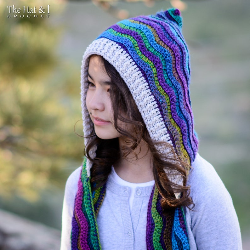 Pu&ograve; includere: Una sciarpa con cappuccio all'uncinetto colorata con un motivo ondulato in tonalit&agrave; di viola, blu, verde e bianco. La sciarpa &egrave; indossata da una persona con i capelli lunghi e castani.