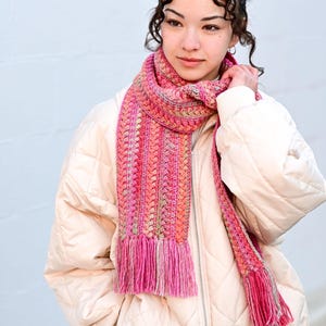 Pu&ograve; includere: Una sciarpa all'uncinetto rosa e arancione con frange. La sciarpa &egrave; indossata da una persona che indossa una giacca trapuntata color crema.