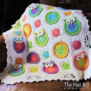 May include: A white crochet baby blanket with colorful owl appliques and circles. The blanket has a scalloped edge and is made with a variety of colors, including pink, blue, green, yellow, and orange. The owls have black eyes and beaks. The text "The Hat & I Crochet" is visible in the bottom right corner of the image.