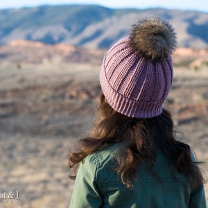 Pu&ograve; includere: Un berretto lavorato a maglia color malva con un pon pon in pelliccia sintetica. Il berretto &egrave; indossato da una persona con i capelli lunghi e castani, in piedi davanti a un paesaggio desertico.