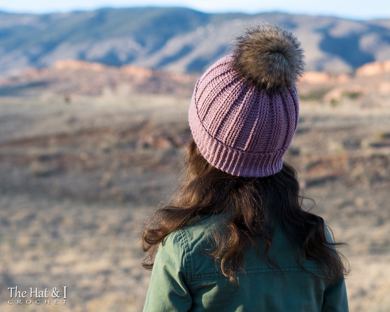 Pu&ograve; includere: Un berretto lavorato a maglia color malva con un pon pon in pelliccia sintetica. Il berretto &egrave; indossato da una persona con i capelli lunghi e castani, in piedi davanti a un paesaggio desertico.