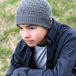 May include: A grey crocheted beanie with a textured woven pattern. The beanie is being worn by a person with dark hair and brown eyes.