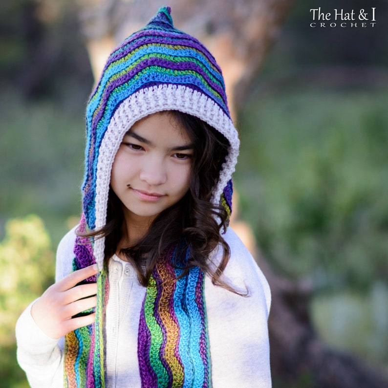 Pu&ograve; includere: Una ragazza indossa una sciarpa con cappuccio all'uncinetto a righe colorate con un bordo all'uncinetto bianco. La sciarpa &egrave; realizzata con una variet&agrave; di colori, tra cui viola, blu, verde e giallo.