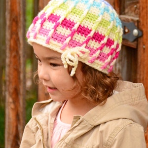 Pu&ograve; includere: Una persona giovane indossa un cappello all'uncinetto colorato con un fiocco bianco. Il cappello ha strisce rosa, verdi, blu e bianche.