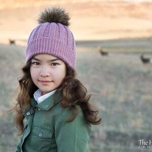 Pu&ograve; includere: Una giovane persona che indossa un cappello di lana rosa con un pon pon soffice. Il cappello ha una trama a coste ed &egrave; indossato su capelli lunghi e castani. La persona indossa una giacca verde e guarda la fotocamera.