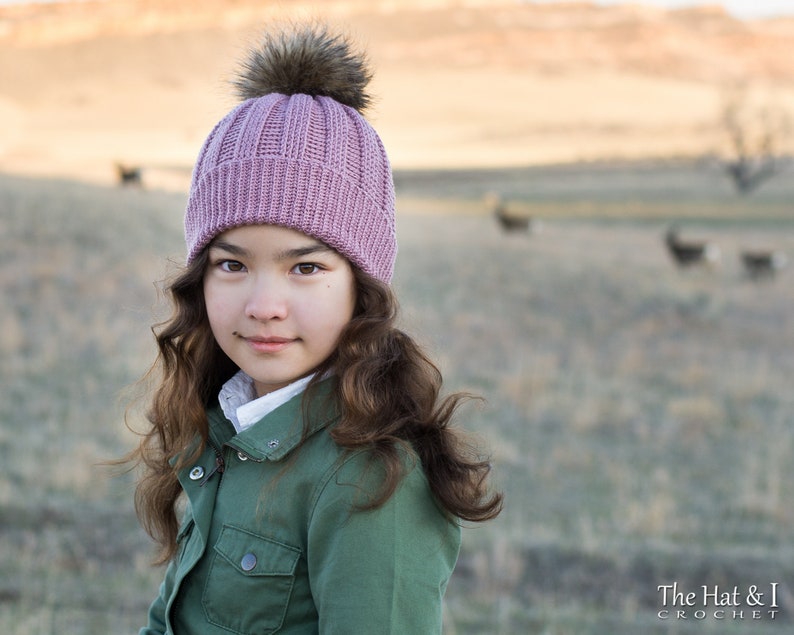 Pu&ograve; includere: Una giovane persona che indossa un cappello di lana rosa con un pon pon soffice. Il cappello ha una trama a coste ed &egrave; indossato su capelli lunghi e castani. La persona indossa una giacca verde e guarda la fotocamera.