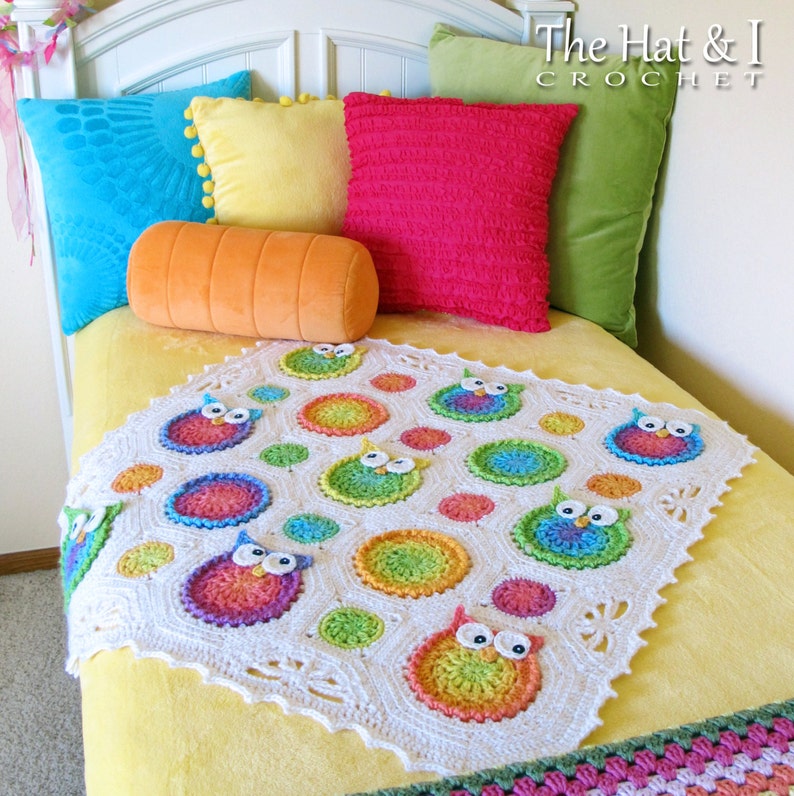 May include: A white crocheted baby blanket with a colorful owl design. The blanket features a repeating pattern of owls and circles in shades of pink, blue, green, and yellow. The blanket is on a yellow bed with pillows in various colors.