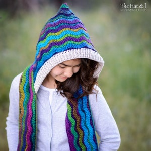 Pu&ograve; includere: Una sciarpa con cappuccio all'uncinetto colorata con un motivo ondulato in tonalit&agrave; di blu, verde, viola e marrone. La sciarpa &egrave; indossata da una persona con i capelli lunghi e castani.