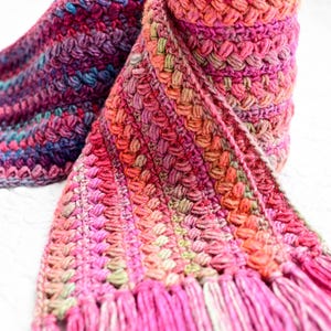 Pu&ograve; includere: Una sciarpa all'uncinetto rosa, verde e viola con frange. La sciarpa &egrave; arrotolata e annodata.