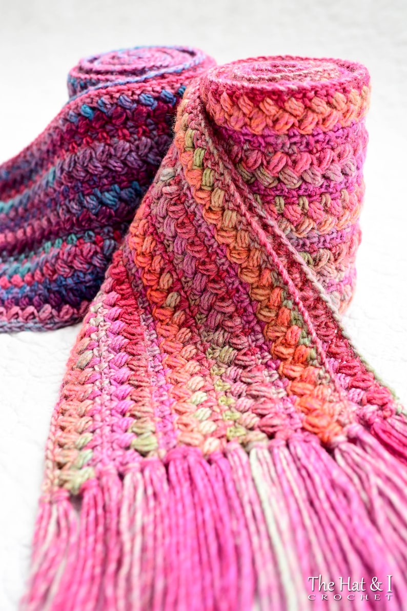 Pu&ograve; includere: Una sciarpa all'uncinetto rosa, verde e viola con frange. La sciarpa &egrave; arrotolata e annodata.