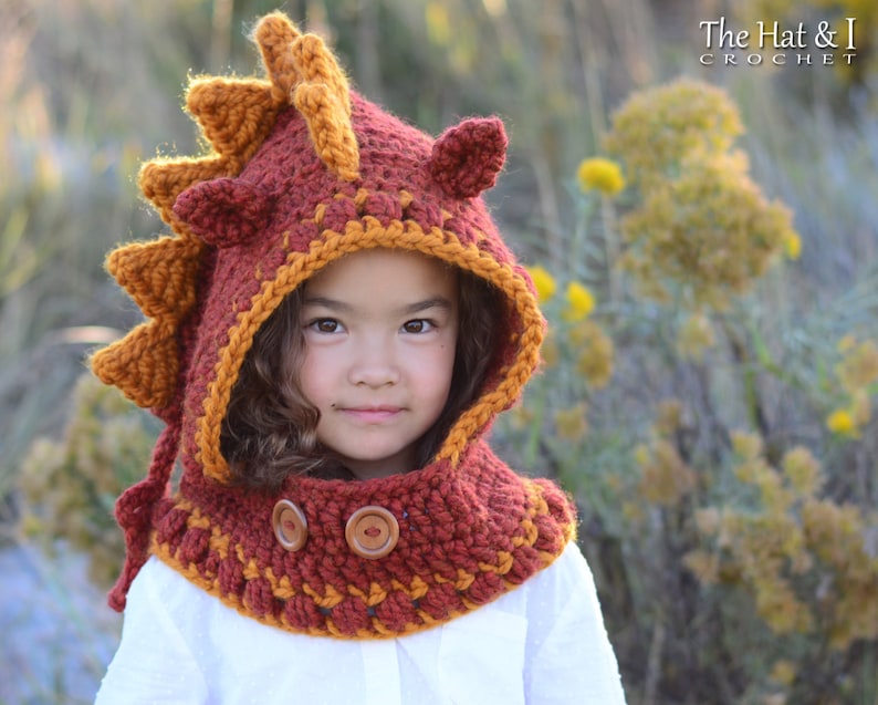 May include: A child wearing a crocheted dragon hooded scarf in shades of orange and brown. The scarf has a large, spiky dragon head with ears and a long tail. The scarf is decorated with brown buttons.