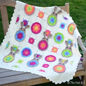 Puede incluir: Una manta de bebé de crochet blanco con un diseño de conejito y flores de colores. La manta presenta una variedad de colores brillantes, incluyendo rosa, azul, verde, amarillo y naranja.