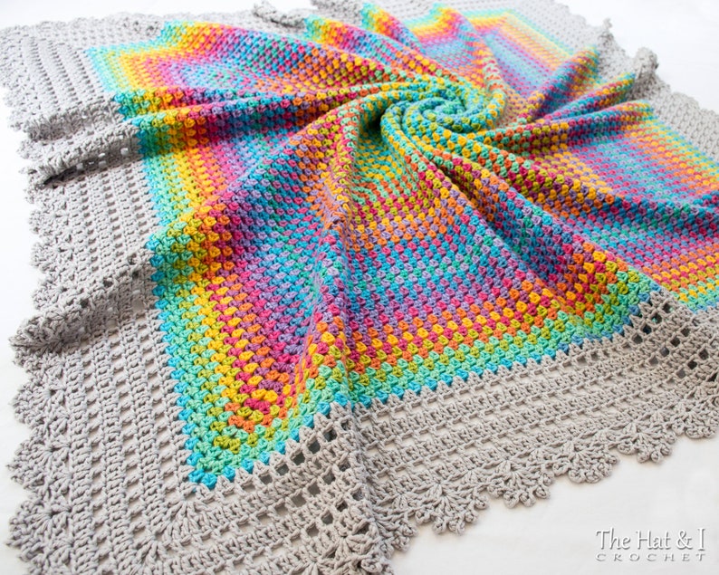 May include: A crocheted blanket with a gray border and a rainbow colored center. The blanket is made with a textured stitch and has a scalloped edge. The text 'The Hat & I CROCHET' is visible in the bottom right corner of the image.