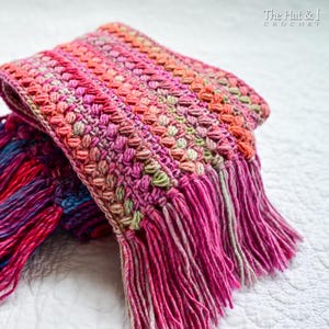Pu&ograve; includere: Una sciarpa all'uncinetto rosa, verde e arancione con frange. La sciarpa &egrave; piegata a met&agrave; e presenta un motivo testurizzato.