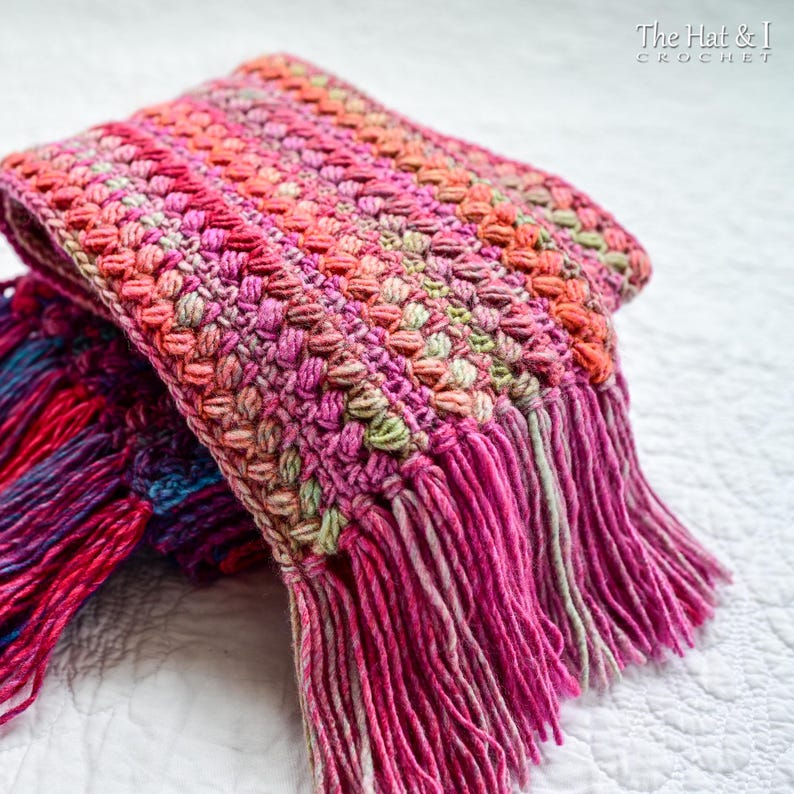 Pu&ograve; includere: Una sciarpa all'uncinetto rosa, verde e arancione con frange. La sciarpa &egrave; piegata a met&agrave; e presenta un motivo testurizzato.