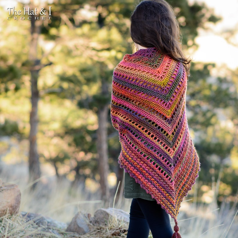Puede incluir: Un chal de ganchillo colorido con un fleco de borlas. El chal est&aacute; hecho con una variedad de colores, incluyendo rosa, naranja, amarillo, verde y morado. El chal est&aacute; drapeado sobre los hombros de una persona de pie en un entorno boscoso.