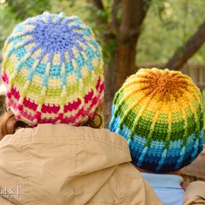 Pu&ograve; includere: Due bambini indossano cappelli all'uncinetto colorati. I cappelli sono realizzati con un arcobaleno di colori, tra cui rosso, arancione, giallo, verde, blu e viola. I cappelli sono all'uncinetto in un motivo circolare, con un design a stella nella parte superiore.