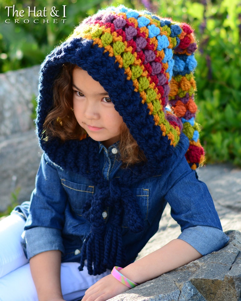 Op de afbeelding: Een kind draagt ​​een blauwe en veelkleurige gebreide capuchon met een lange kwast. De muts heeft een kleurrijk bobbelsteekpatroon. De muts heet "The Hat & I Crochet".