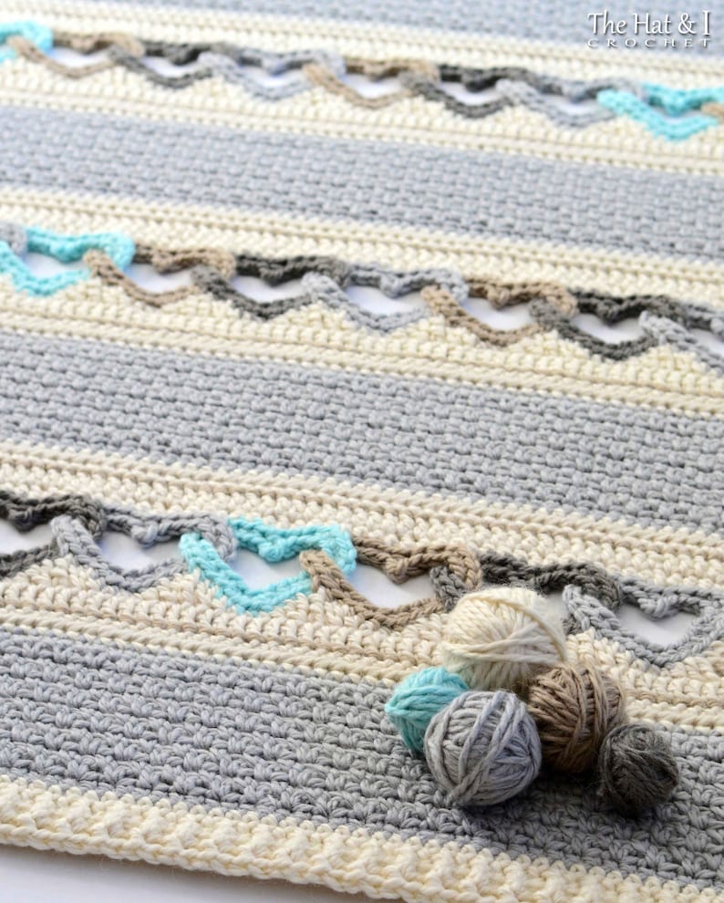 May include: A crocheted blanket with a striped pattern in shades of gray, beige, and blue. The blanket has a unique design with a series of connected hearts. There are four balls of yarn in different colors, including gray, beige, and blue, lying on the blanket.