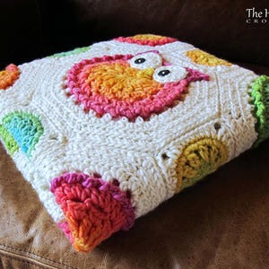 May include: A white crochet baby blanket with a colorful owl design in the center. The blanket is made with a variety of colors, including pink, blue, green, yellow, and orange. The owl has large, black eyes and a pink beak. The blanket is folded in half and is sitting on a brown leather couch.