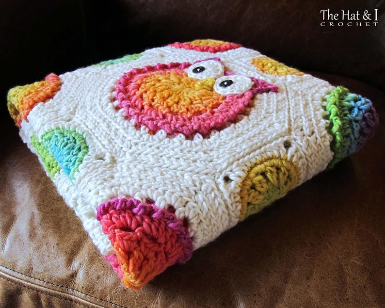 May include: A white crochet baby blanket with a colorful owl design in the center. The blanket is made with a variety of colors, including pink, blue, green, yellow, and orange. The owl has large, black eyes and a pink beak. The blanket is folded in half and is sitting on a brown leather couch.