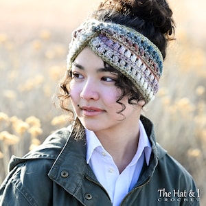 Può includere: Una donna indossa una fascia per capelli all'uncinetto multicolore con un design a croce. La fascia è realizzata con una varietà di tonalità di marrone, verde, blu e viola. La donna indossa una camicia bianca e una giacca verde. The Hat & I Crochet è stampato nella parte inferiore dell'immagine.