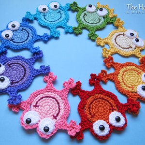 May include: A rainbow of crocheted frog appliques with black button eyes. The frogs are arranged in a circle on a blue background.