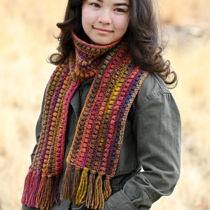 Puede incluir: Una persona lleva una larga bufanda de ganchillo multicolor con borlas. La bufanda tiene un dise&ntilde;o texturizado y abultado y presenta tonos de rojo, naranja, amarillo, marr&oacute;n y morado.