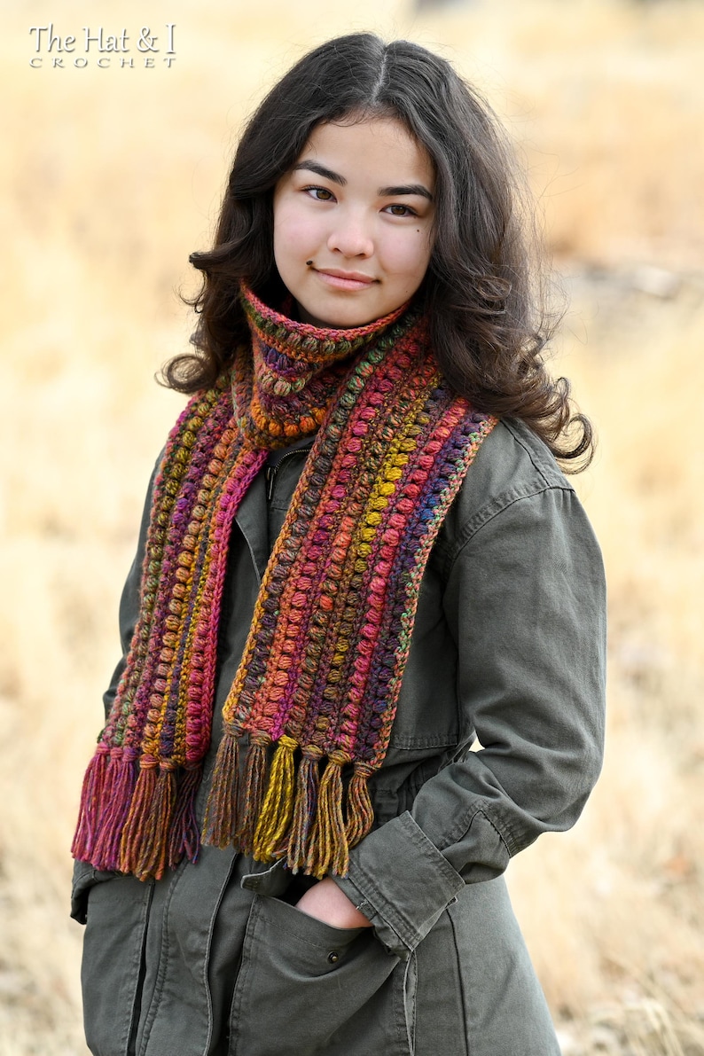 Puede incluir: Una persona lleva una larga bufanda de ganchillo multicolor con borlas. La bufanda tiene un dise&ntilde;o texturizado y abultado y presenta tonos de rojo, naranja, amarillo, marr&oacute;n y morado.