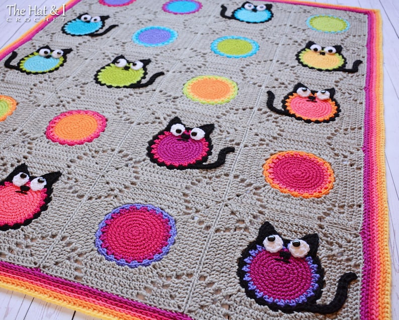 May include: A gray crochet blanket with a colorful cat design. The blanket features a repeating pattern of circles and cats with large eyes. The blanket has a multi-colored border.