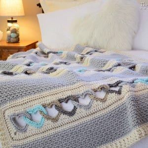 May include: A crocheted throw blanket with a heart pattern in shades of gray, beige, and light blue. The blanket is draped over a bed with white bedding.