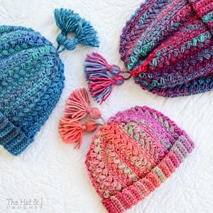 May include: Three crocheted hats with colourful yarn and tassels. The hats are in shades of blue, pink and purple. The hats are on a white surface. The text "The Hat & I Crochet" is in the bottom left corner of the image.