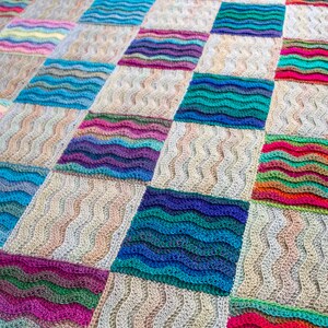 Pode incluir: Uma manta de croch&ecirc; colorida com um padr&atilde;o ondulado. A manta &eacute; composta por quadrados de diferentes cores, incluindo azul, verde, rosa e roxo. A manta &eacute; feita com um fio macio e tem um aspecto texturizado.
