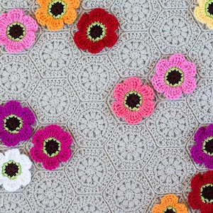 May include: A crocheted blanket with a hexagonal pattern in a light gray color. The blanket is decorated with various crocheted flowers in different colors, including pink, red, orange, white, and purple.