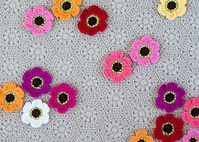 May include: A crocheted blanket with a hexagonal pattern in a light gray color. The blanket is decorated with various crocheted flowers in different colors, including pink, red, orange, white, and purple.