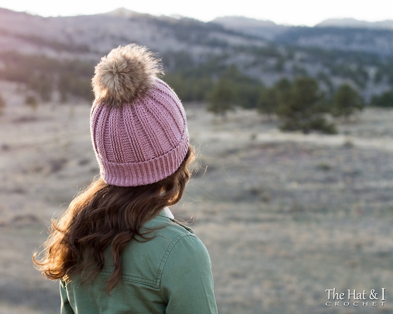 May include: A pink crocheted beanie with a brown faux fur pom pom. The beanie is worn by a person standing in a field with a mountain range in the background.