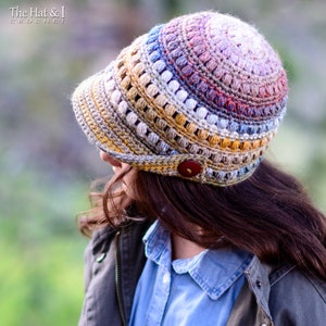 May include: A crocheted hat with a colourful ombre design and a button detail. The hat has a wide brim and is made with a variety of stitches. The hat is worn by a person with long brown hair.