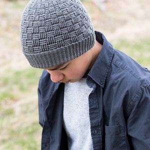 May include: A grey crocheted beanie with a woven pattern. The beanie is being worn by a person with a blue shirt.