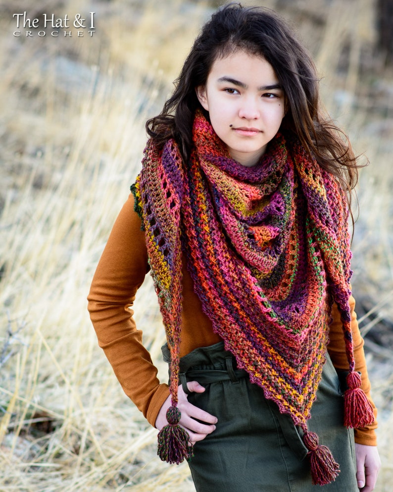 Puede incluir: Una mujer lleva una chalina de ganchillo de colores con borlas. La chalina es de forma triangular con un degradado de colores, incluyendo rojo, naranja, amarillo, verde y morado. La mujer lleva una camisa de manga larga marr&oacute;n y una falda verde.
