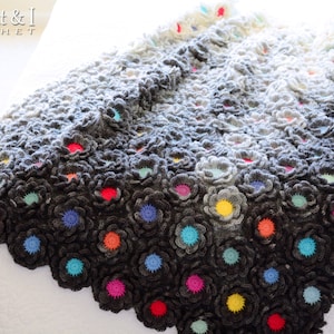 Puede incluir: Una manta de ganchillo con un degradado de hilo gris, que presenta un patr&oacute;n repetitivo de flores con centros coloridos. La manta est&aacute; hecha con un hilo suave y texturizado y tiene un aspecto delicado y femenino.