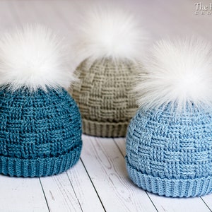 May include: Three crocheted hats with white faux fur pom poms. The hats are in shades of blue and grey. The hats are on a white wooden surface.