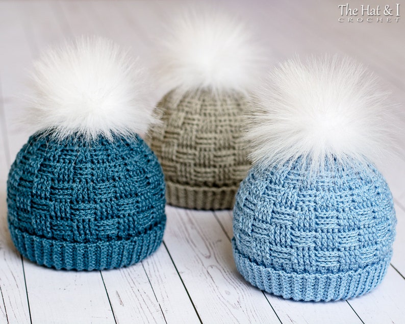 May include: Three crocheted hats with white faux fur pom poms. The hats are in shades of blue and grey. The hats are on a white wooden surface.
