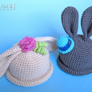 Puede incluir: Dos gorros de conejo de ganchillo, uno en beige con una flor rosa y hojas verdes, y el otro en gris con un detalle de ojo azul y blanco. Los gorros están sobre un fondo azul claro.
